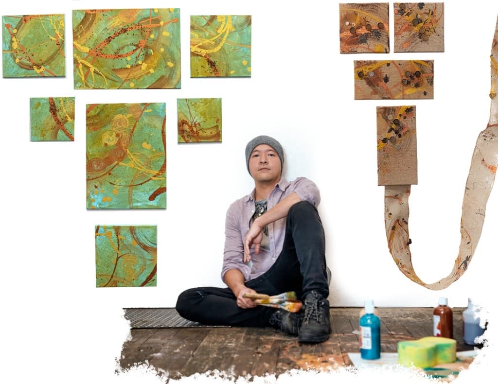 Artist William Ho sitting on a paint-stained floor beneath his abstract canvases