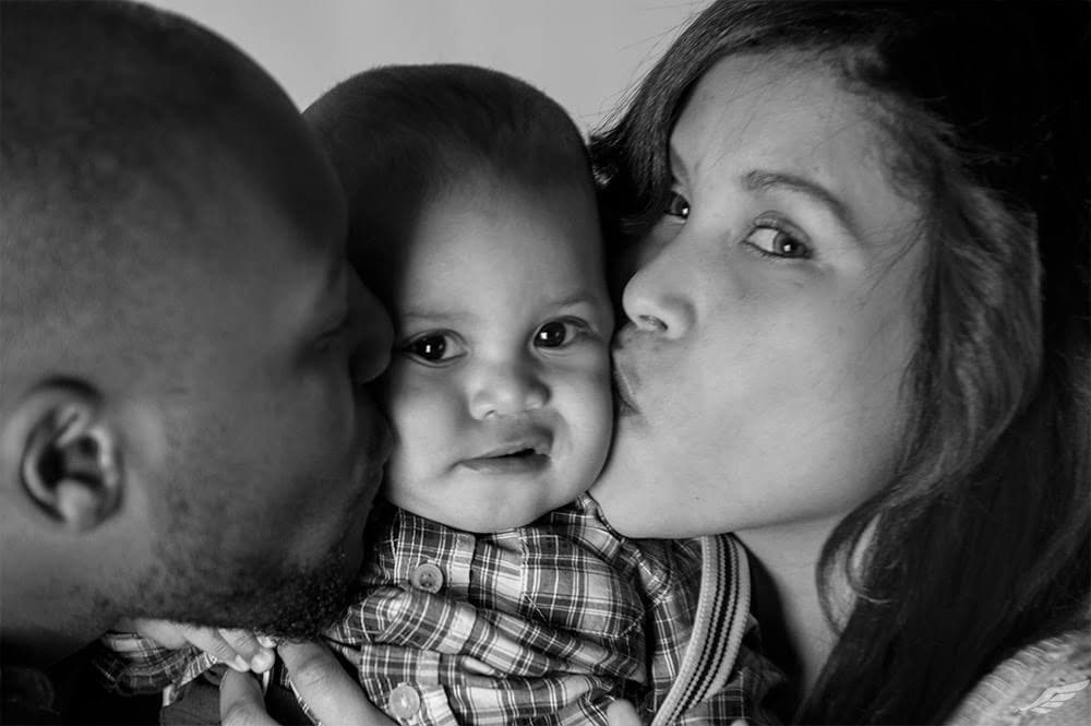 Black and white photo of two parents kissing their baby on each cheek